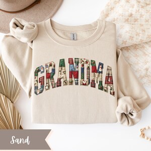 May include: A sand-colored sweatshirt with the word "GRANDMA" in patchwork letters. The letters are made of various plaid and patterned fabrics in red, green, blue, and white. The sweatshirt is folded neatly on a white surface.