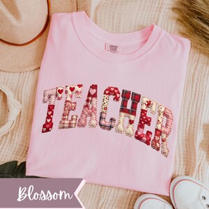 May include: A pale pink t-shirt featuring the word "TEACHER" in appliqu&eacute;d fabric letters. The letters incorporate heart and plaid patterns. The shirt is a Comfort Colors brand.