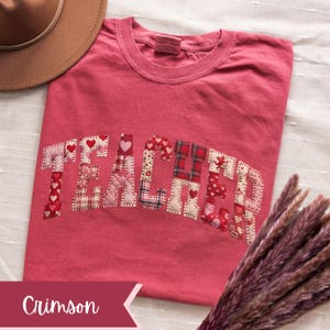 May include: Crimson t-shirt featuring the word "TEACHER" in patchwork. The patchwork incorporates heart and plaid patterns. A brown hat is partially visible in the background. The word "Crimson" is written on a banner.