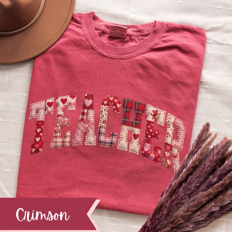 May include: Crimson t-shirt featuring the word "TEACHER" in patchwork. The patchwork incorporates heart and plaid patterns. A brown hat is partially visible in the background. The word "Crimson" is written on a banner.