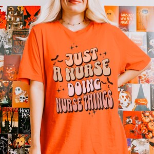 May include: Orange t-shirt with the text "JUST A NURSE DOING NURSE THINGS" in a retro font. The shirt has a V-neck and short sleeves. The background features Halloween-themed posters.