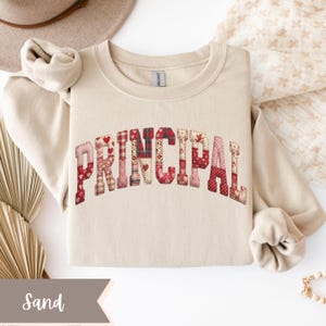 May include: A sand-colored sweatshirt with the word "PRINCIPAL" across the chest. The letters are made of a patchwork design with red, pink, and white patterns, including hearts and plaid. The sweatshirt has a crew neck and long sleeves.