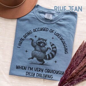 May include: Blue jean colored t-shirt with a raccoon graphic. The raccoon is dancing with its arms up and tail curled. Text on the shirt reads "I hate being accused of lollygagging when I'm very obviously dilly dallying."