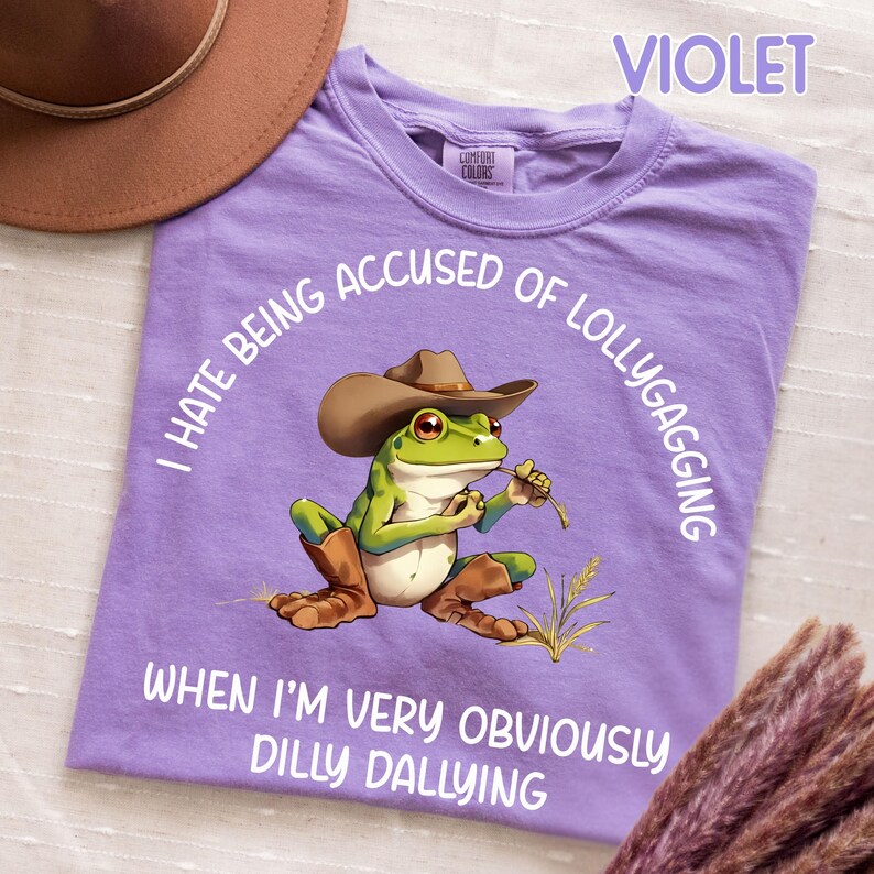 May include: Violet Comfort Colors t-shirt with a cartoon frog wearing a cowboy hat and boots. The frog is holding a piece of grass. Text on the shirt reads "I HATE BEING ACCUSED OF LOLLYGAGGING WHEN I'M VERY OBVIOUSLY DILLY DALLYING."
