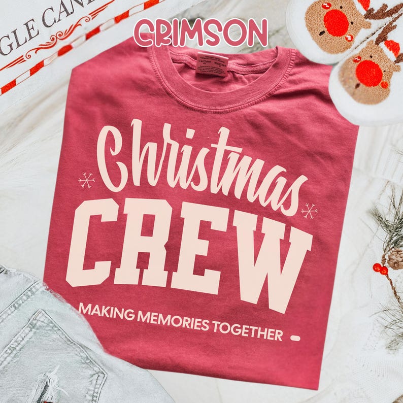May include: Crimson red t-shirt with the words "Christmas CREW" in white block letters. The phrase "MAKING MEMORIES TOGETHER" is printed below. The shirt is displayed with reindeer slippers and festive decorations.