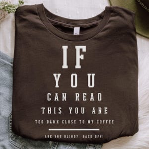 May include: A dark brown t-shirt with white text. The text reads "IF YOU CAN READ THIS YOU ARE TOO DAMN CLOSE TO MY COFFEE. ARE YOU BLIND? BACK OFF!" The shirt is folded and lies on a light-colored surface.