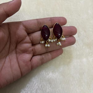 May include: A pair of gold-toned earrings featuring a dark red, oval-shaped stone. Each earring has a cluster of small, white pearls dangling from the bottom. The earrings are displayed on a light-colored surface.