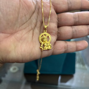May include: A gold-colored pendant necklace featuring a detailed figure within a sunburst design. The pendant hangs from a delicate chain. The necklace is held in a hand, with a teal-colored box in the background.