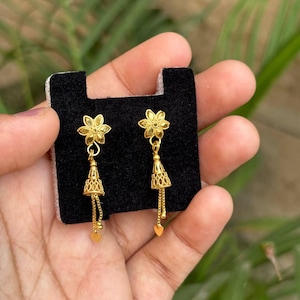 May include: A pair of gold-colored earrings. Each earring features a flower-shaped top, a bell-shaped middle section, and a heart-shaped charm at the bottom. The earrings are displayed on a black surface.