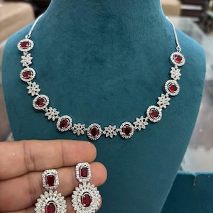 May include: A silver necklace and earring set. The necklace has oval red gemstones set amongst small clear stones. The earrings mirror the design, with a red gemstone centre and a halo of clear stones. The jewellery is displayed on a teal mannequin.