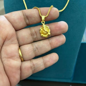 Könnte beinhalten: Eine goldfarbene Halskette mit einem Anhänger, der Ganesh darstellt. Der Anhänger hängt an einer feinen Kette. Die Halskette wird vor einem türkisfarbenen Hintergrund präsentiert.