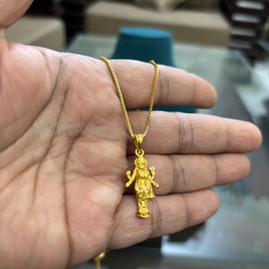 May include: A gold-colored pendant necklace featuring a detailed figure of a deity. The pendant is suspended from a delicate chain. The figure is intricately designed, showcasing fine details. The necklace is held in a hand, suggesting it is being displayed.