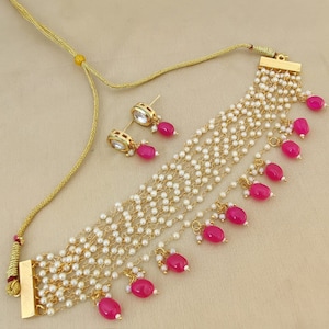 May include: A gold necklace with multiple strands of white pearls and pink gemstone drops. The necklace has a matching pair of earrings with a clear stone center and pink gemstone drops.