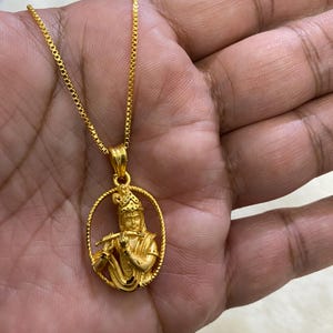 May include: A gold-colored pendant necklace featuring a detailed depiction of a figure playing a flute, set within an oval frame. The pendant hangs from a delicate gold chain, showcasing intricate craftsmanship. The necklace is a piece of jewelry.