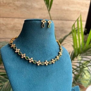 May include: A gold-toned necklace and matching stud earrings set featuring alternating oval-shaped green stones and small clear white stones arranged in a floral pattern. The jewelry is displayed on a teal-colored velvet-textured bust form.