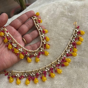 Könnte beinhalten: Ein goldfarbenes Halsketten- und Kopfschmuck-Set mit kleinen weißen Kunstperlen, leuchtend gelben tropfenförmigen Perlen und Büscheln aus winzigen magentafarbenen Perlen. Der Schmuck ist mit klaren, blütenblattförmigen Steinen verziert.