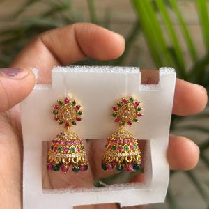 May include: A pair of gold-tone jhumka earrings featuring a bell-shaped design adorned with small, alternating pink and green stones, clear crystals, and dangling beads at the base, attached to a circular stud with a central teardrop-shaped stone.