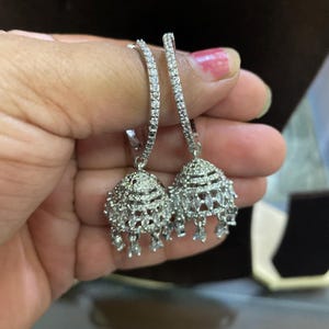May include: Silver hoop earrings with dangling, teardrop-shaped charms. The charms are covered in sparkling white stones.