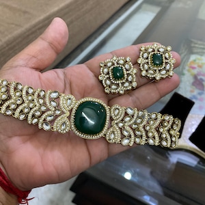 Può includere: Un set di gioielli color oro con un braccialetto e orecchini abbinati. Il braccialetto ha una grande pietra centrale quadrata verde smeraldo, circondata da pietre chiare più piccole. Gli orecchini hanno un design simile. I gioielli sono esposti su una mano.