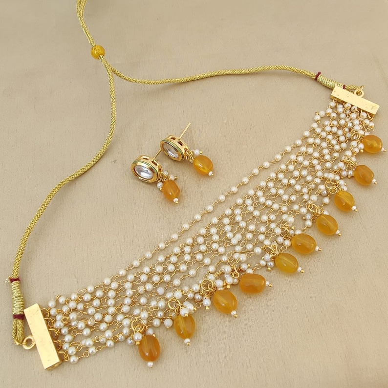May include: A gold necklace with multiple strands of small white beads and yellow teardrop-shaped stones. The necklace has a gold clasp and comes with a matching pair of earrings with a clear stone center and yellow teardrop-shaped stones.