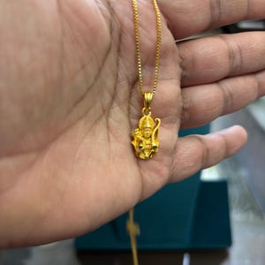 May include: A gold-colored pendant necklace featuring a detailed figure. The pendant is suspended from a delicate chain. The figure has intricate details and is likely made of gold. The necklace is held in a hand.