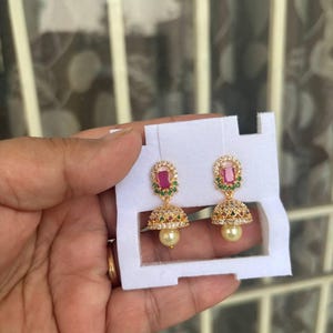 May include: A pair of gold-colored earrings with a rectangular red stone and small green stones at the top. The earrings have a bell-shaped design with a pearl-like bead at the bottom and are set on a white card.