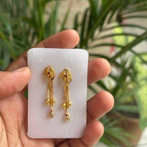 May include: A pair of gold dangle earrings featuring a marquise-shaped top with a small red stone and clear accent stones. Three delicate gold chains hang from each top, ending in a small gold bead and a teardrop-shaped charm with a red stone.