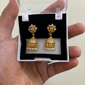 May include: A pair of gold earrings with green and red stones, and white pearls hanging from the bottom. The earrings are in a clear plastic box.