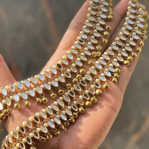 May include: Gold-toned anklets with intricate detailing. Each anklet features a series of mirrored teardrop-shaped accents, small pearls, and dangling gold beads. The jewelry is displayed against a blurred background, highlighting the anklets' design.