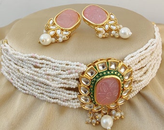 May include: A white pearl choker necklace with a pink stone and gold accents. The necklace has multiple strands of pearls and a large, ornate pendant with a pink stone in the center. The pendant is surrounded by smaller stones and gold details. The necklace comes with a pair of matching earrings with pink stones and gold accents.