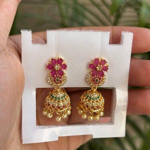 May include: A pair of gold-tone jhumka earrings featuring a flower-shaped stud with pink stones, a crescent-shaped accent with green and clear stones, and a bell-shaped drop adorned with pink, green, and clear stones, finished with small dangling beads.