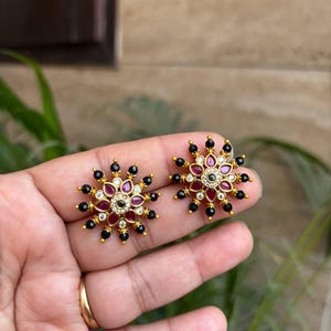 May include: A pair of gold-toned stud earrings. Each earring features a floral design with red, black, and clear gemstones. The earrings are held in a hand, showcasing their intricate details and craftsmanship.