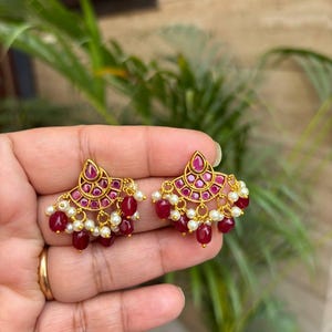 May include: Gold-toned earrings with a fan-shaped design. The earrings feature a cluster of small, round, red gemstones and dangling red beads. Small white pearls are also incorporated into the design, adding a touch of elegance.