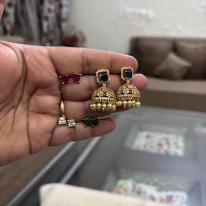 May include: A collection of gold-toned earrings, including large jhumka-style earrings with blue square stones and pearl accents. Other earrings include small ruby, emerald, and diamond-like studs, and a small hoop earring.