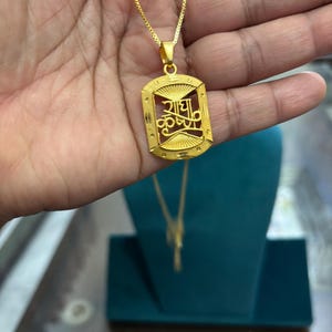 May include: A gold-colored pendant necklace with a rectangular pendant. The pendant features an intricate design with text in a script font. The necklace is displayed on a teal jewelry stand.