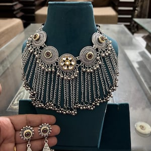 May include: A silver-toned necklace featuring circular medallions with central red and white stone accents, adorned with multiple cascading silver chains and small spherical beads. The set includes matching drop earrings with similar circular tops.
