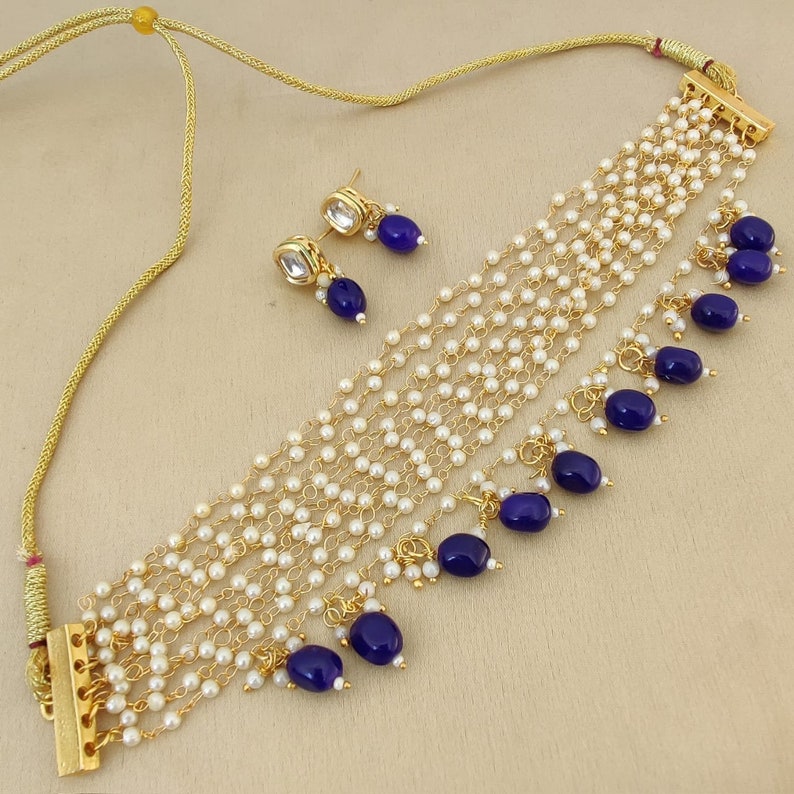 May include: A gold necklace with multiple strands of white pearls and blue gemstone accents. The necklace has a clasp closure and comes with a matching pair of earrings.