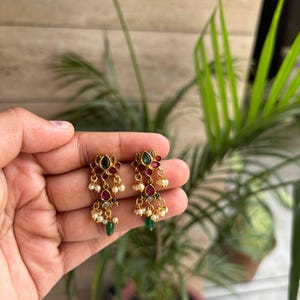 May include: A pair of gold-toned drop earrings featuring a central lotus-shaped design with inset red and green stones. Each earring is accented with small dangling white beads and a single green teardrop-shaped stone at the bottom base.
