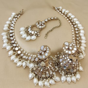 May include: A gold and white necklace and earrings set with pearl accents. The necklace has multiple strands of small mirrors and pearls. The earrings are large and feature a round design with mirrors and pearls.