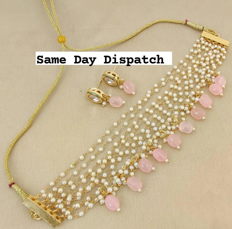 May include: A gold necklace with multiple strands of pearls and pink stone accents. The necklace has a clasp closure and comes with a matching pair of earrings with pink stones.
