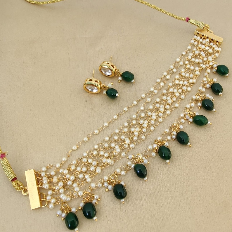 May include: A gold necklace with multiple strands of white pearls and green gemstone accents. The necklace has a clasp closure and comes with matching earrings.