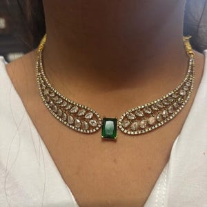 以下が含まれることがあります： 中央にエメラルドカットの緑色の宝石をあしらったゴールドトーンのネックレス。ネックレスは、クリアで輝く石が多数あしらわれ、繊細で対称的なパターンに配置されています。白い背景にデザインされています。