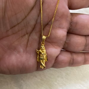 May include: A gold-colored pendant necklace featuring a detailed figure. The pendant is suspended from a delicate chain. The figure appears to be a religious or mythological character. The necklace is displayed against a neutral background.