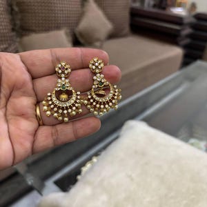 May include: Gold-toned chandelier earrings with intricate detailing. Each earring features a floral stud, a crescent-shaped design with small pearls, and a central bell-shaped element. The earrings are adorned with clear and colored gemstones.