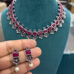 May include: A silver necklace and earring set with rectangular, ruby-red gemstones. The necklace and earrings have a floral design, with a pearl drop on the earrings. The jewellery is displayed on a teal mannequin.
