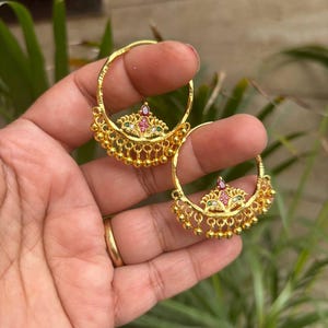 May include: Gold-toned hoop earrings with intricate detailing. Each earring features a floral design with red, green, and white gemstones, and small dangling gold beads. The earrings are held up against a green leafy background.