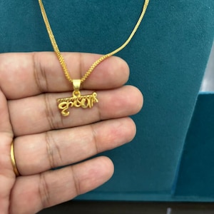 May include: A gold-colored necklace with a pendant featuring script. The pendant is suspended from a delicate chain. The necklace is displayed against a teal background.