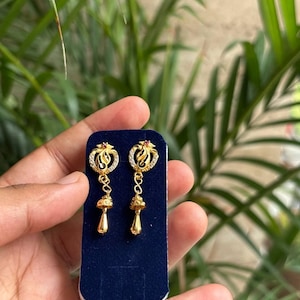 May include: A pair of gold drop earrings featuring a circular top section with small clear stones and a tiny red accent, connected to a dangling gold chain with a bell-shaped charm and a smooth, teardrop-shaped gold pendant at the bottom.
