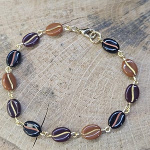 May include: A gold-toned chain bracelet featuring oval-shaped beads in shades of brown, black, and purple. Each bead has a gold-colored stripe. The bracelet is secured with a lobster clasp, and the beads resemble coffee beans.