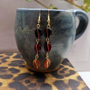 May include: A pair of dangle earrings with gold-colored hooks. Each earring features three beads in shades of brown and amber, with a dark red stripe. The earrings are displayed against a blue ceramic mug and a leopard print surface.